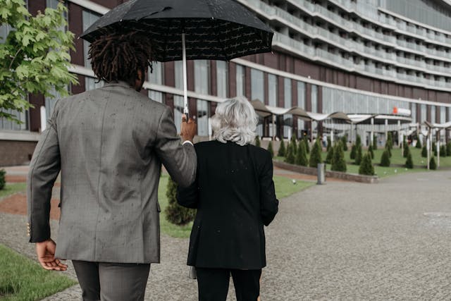 man in brown suit holding black umbrella at the back of a senior woman walking in black blazer